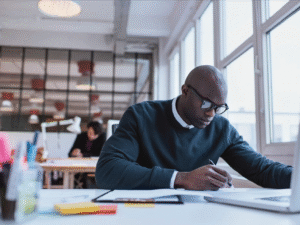 Focused professional writing notes at a desk with a laptop and office materials